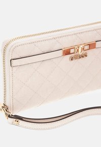 Porte-monnaie Guess, en faux cuir crème, motif matelassé, quincaillerie dorée, fermeture zippée, sangle de poignet amovible, accent logo sur le devant.