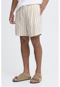 Shorts en lin rayés beige et blanc avec une taille élastique, des poches latérales et une coupe décontractée, associés à des sandales à glisser marron clair.