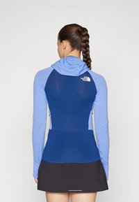 Top sportiv albastru cu mâneci lungi și glugă, având un design texturat și logo pe spate. Purtați cu un skort negru.