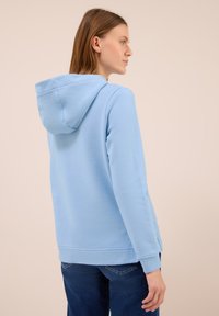 Donna di profilo che indossa un felpa azzurra chiara e jeans blu scuro, su uno sfondo beige uniforme.