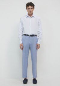 Un homme se tient contre un fond blanc, portant une chemise blanche à manches longues boutonnée, un pantalon bleu clair avec une ceinture noire, et des mocassins noirs.