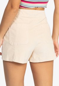 Shorts en velours côtelé beige avec une taille haute, deux poches arrière et une coupe droite, mettant en avant une texture douce et un design ajusté.