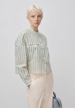 Junge Person mit kurzen blonden Haaren trägt ein grün-weiß vertikal gestreiftes Hemd mit Knöpfen und hellbeige Hosen, hält eine Tasche in der Hand.