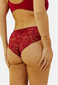 Culotte en dentelle rouge profond, ornée de motifs floraux et de bords festonnés. Texture douce avec un design ajusté. Bracelet en or au poignet.