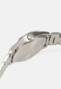 Montre en acier inoxydable argenté avec un boîtier rond poli, une couronne texturée et un bracelet à maillons. Le dos présente des détails gravés.