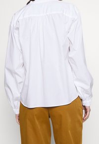 Vit bomullsskjorta med långa, sammanknipna ärmar och rundad nederkant, ihop med bruna vida byxor. Enkelt design, inga synliga mönster.