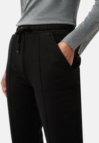 Zwarte joggingbroek met elastische tailleband, zakken en trekkoord. Zachte textuur en contrasterende gestikte lijnen. Grijze lange mouwen top op de achtergrond.