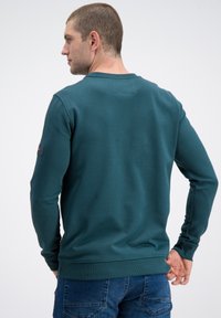 Bluza turkusowa z okrągłym dekoltem, ściągaczami na mankietach i na dole, gładką fakturą oraz naszywką z logo na lewym rękawie. Luźny krój.