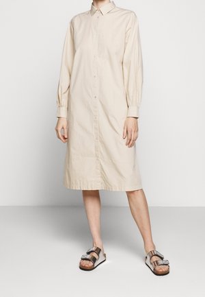 Personne portant une robe-chemise longue beige à boutons avec manches longues et des sandales argentées métalliques à deux brides.