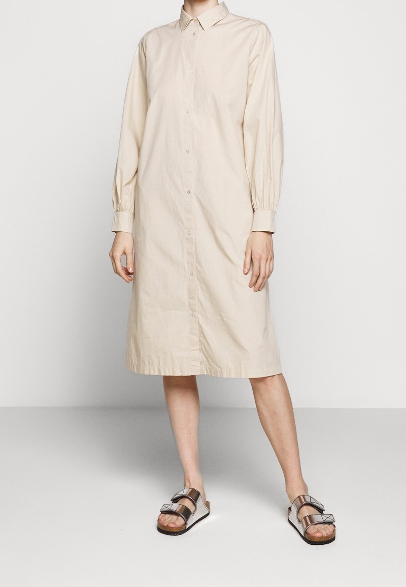 Personne portant une robe-chemise longue beige à boutons avec manches longues et des sandales argentées métalliques à deux brides.