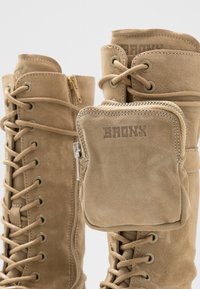 Botas de tobillo de ante en tono beige con cordones en la parte delantera, ojales de metal y cremallera lateral. Presenta un pequeño compartimento con cremallera sujeto a la parte superior.