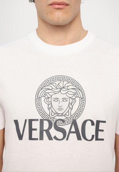 Hvid bomulds t-shirt med et centreret sort græsk nøgle-design og Medusa-hoved samt fed "VERSACE" tekst nedenfor i sort.