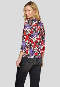 Bluse mit floralem Muster in Rot, Lila und Schwarz. Dreiviertelärmel und eine lockere Passform. Kombiniert mit einer dunklen Jeans.