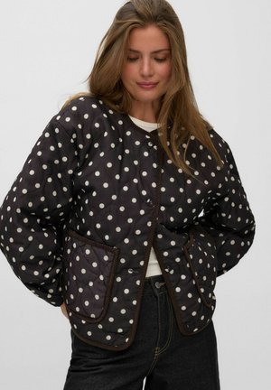 Femme portant une veste matelassée noire à pois blancs avec de grandes poches avant, associée à un jean foncé et un haut blanc en dessous.