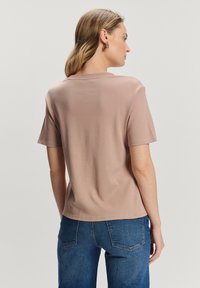 Kobieta z jasnobrązowymi włosami, ubrana w beżową koszulę z krótkim rękawem i niebieskie dżinsy, pokazana od tyłu na gładkim tle.