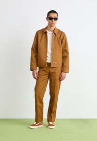 Tan jacket and pants made of thick fabric, white shirt underneath, with sunglasses and brown suede sneakers, standing on a green surface.