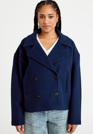 Kobieta z zaplecionymi włosami, ubrana w dwurzędowy, ciemnoniebieski wełniany płaszcz i jasnoniebieskie jeansy, stojąca na jednolitym białym tle.