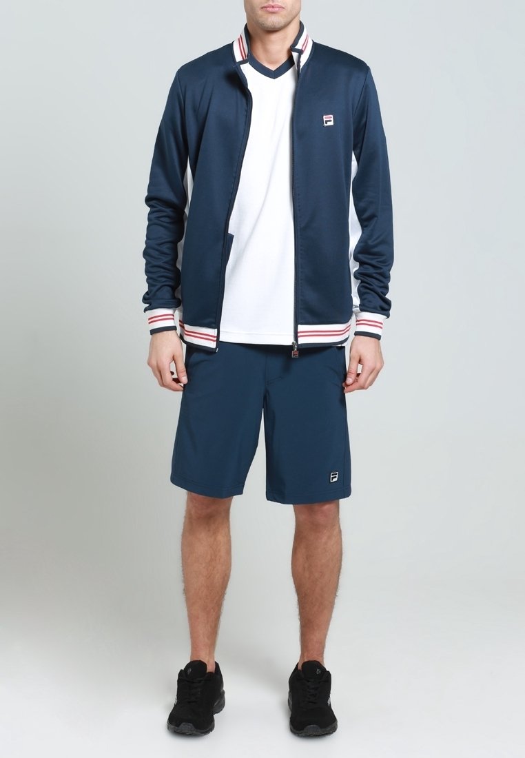 Veste zippée de sport marine avec des accents rayés rouge et blanc, chemise blanche en dessous, et shorts marins. Des chaussures noires complètent la tenue.