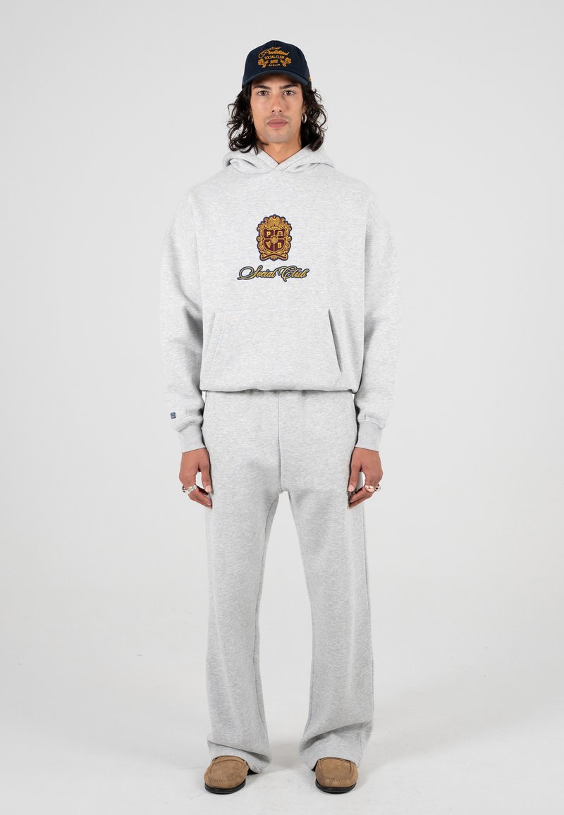 Felpa grigia con cappuccio e tasca frontale, caratterizzata da un logo stampato colorato. Abbinata a pantaloni della tuta grigi e scarpe slip-on marroni.