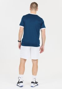 Marinblå sporttröja med vita accenter, ihop med vita shorts. Har rund hals och strukturerad tyg. Svarta träningsskor med vita strumpor.