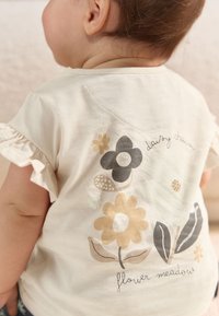 Cremefarbenes Baumwoll-T-Shirt mit gerafften Ärmeln. Mit floralen Designs in Schwarz, Gelb und Braun. Text lautet "daisy chain" und "flower meadow".