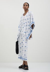 Set di tunica e pantaloni bianchi con motivi floreali blu, vestibilità rilassata, tessuto testurizzato, abbinato a una borsa intrecciata nera e sandali con zeppa.