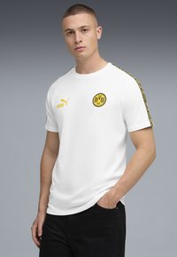 Ung mand med kort hår iført en hvid Borussia Dortmund fodbold T-shirt med gult logo og mønstrede ærmestriber, poserer med den ene hånd i lommen.