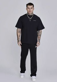 T-shirt noir à manches courtes avec col rond et logo blanc, associé à un pantalon noir, caractérisé par une coupe ajustée et un détail de logo sur le côté.