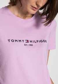 Ljuslila bomullströja med "TOMMY HILFIGER"-logotyp i röd, vit och blå accent. Korta ärmar och rundad halsringning.