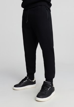 Pantaloni della tuta neri con un design affusolato, caviglie elastiche e tasche laterali. Indossati con sneakers nere e bianche con suole testurizzate.