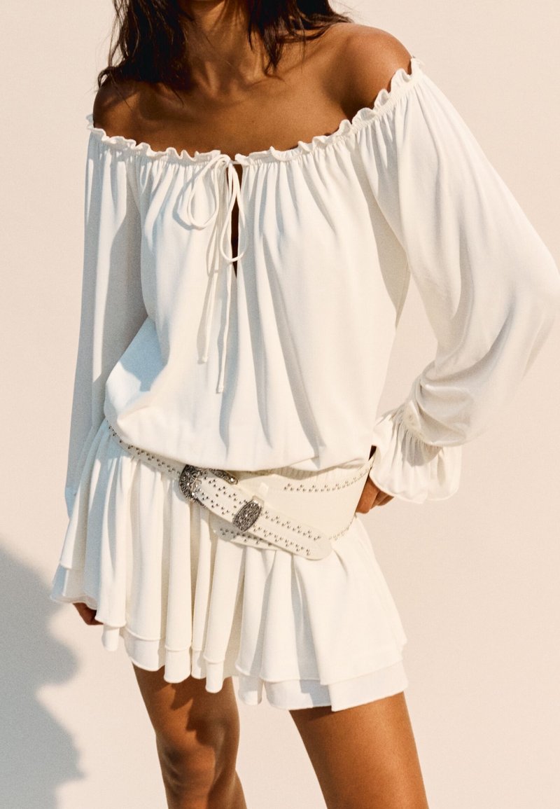 Bílá šaty s odhalenými rameny, s volánkovým výstřihem, detailem na šňůrku, dlouhými rukávy a vrstveným lemem. Doplněné o opasek se cvočky.