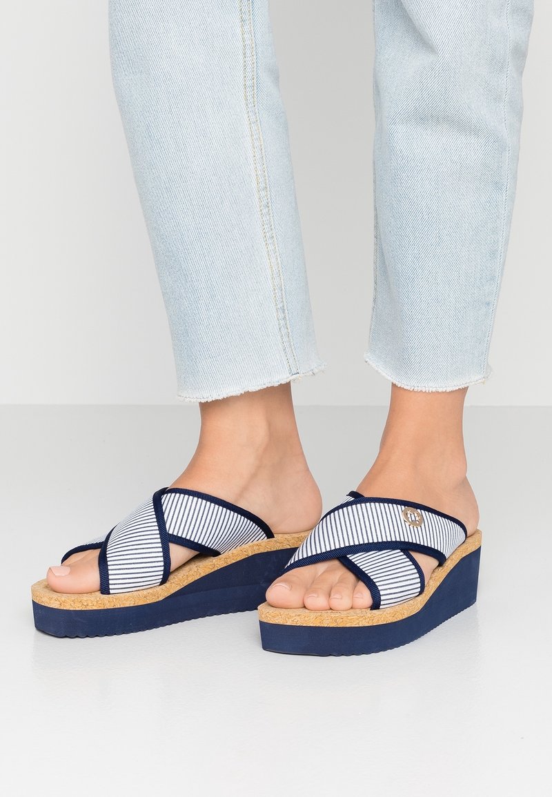 Mules compensées à rayures marines avec une plateforme en liège. Dotées de brides en tissu croisées et d'un petit bouton d'accent sur le côté.