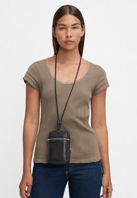 Frau in einem taupefarbenen Kurzarmoberteil und dunklen Jeans, mit einer schwarzen Lederpouch, die an einer langen Halskette um ihren Hals hängt.