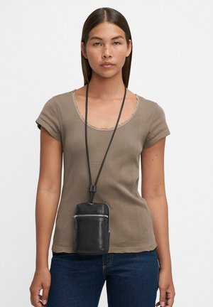 Frau in einem taupefarbenen Kurzarmoberteil und dunklen Jeans, mit einer schwarzen Lederpouch, die an einer langen Halskette um ihren Hals hängt.