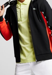 Svart och orange dragkedjetröja med mjuk textur, prydd med en grön logotyp och handmönster på ärmarna, bärs över en ljusgrön polo.