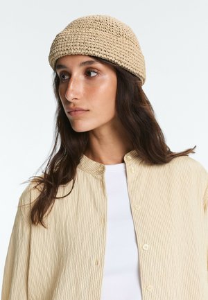 Giovane donna con lunghi capelli scuri che indossa un cappello beige intrecciato e una camicia a righe beige chiaro sopra una maglietta bianca, guarda verso sinistra su uno sfondo neutro.