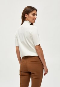 Blusa blanca de manga corta con un cuello alto, combinada con pantalones marrones ajustados. La tela parece suave con una sutil textura, sin patrones visibles.