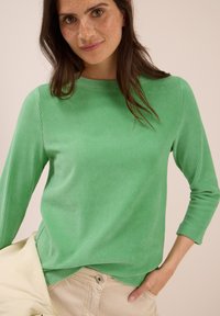 Eine Frau mit langen, dunklen Haaren und Sommersprossen trägt ein grünes, geripptes Langarmoberteil, beige Hosen und hält eine hellbeige Jacke an ihrer Seite.