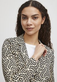 Blazer à motifs en crème et noir, présentant des designs géométriques. Le matériau semble tissé avec une texture lisse. Fermeture à boutons, design à col.