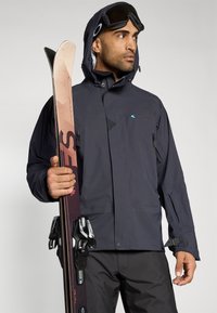 Tamsiai mėlynas slidinėjimo švarkas su vandeniui atsparia medžiaga, užtrauktukais užsegamomis kišenėmis, reguliuojamais rankogaliais ir tinkleliu viduje. Dešinėje rankoje laikomos slidės.