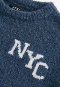Pull en maille bleue floue avec des lettres blanches texturées "NYC" sur le devant et une étiquette noire "NEXT" à l'intérieur du col.