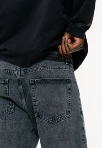 Chemise noire à manches longues rentrée dans un jean en denim gris. Le jean présente une finition texturée et deux poches arrière. Étiquette de la marque visible à la taille.