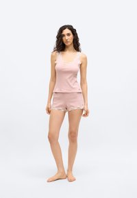 Frau mit lockigem Haar, die ein rosa ärmelloses Oberteil und passende Shorts mit Spitzenbesatz trägt, steht barfuß vor einem schlichten Hintergrund.