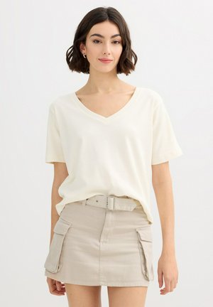 Mujer con cabello corto y oscuro que lleva una camiseta blanca de cuello en V metida en una minifalda beige con cinturón y grandes bolsillos delanteros.