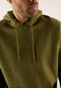 Olivgrüner Hoodie mit Kordelzug, weicher Textur, Fronttasche und lockerer Passform. Der Stoff wirkt warm und bequem.