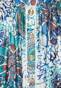 Robe en tissu imprimé avec un motif floral et géométrique vibrant en bleu et vert, ornée de boutons dorés et de détails blancs sur le devant.