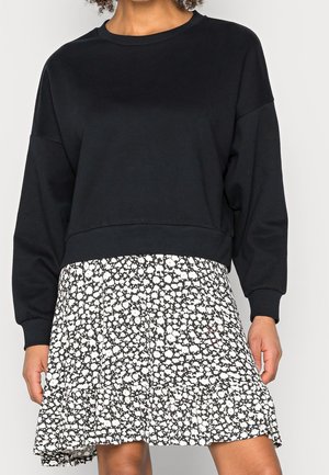 Kobieta ubrana w czarną bluzę z długim rękawem, zestawioną z czarno-białą, wzorzystą spódnicą do kolan.