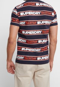 Mørkeblå bomulds t-shirt med vandret hvid og orange tekst "SUPERDRY TRACKSTARS". Klassisk pasform med korte ærmer og rund hals.