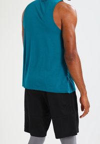 Uomo che indossa una maglietta atletica senza maniche di colore teal, pantaloni neri e leggings grigi, visto da dietro su uno sfondo neutro.