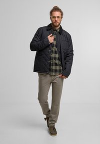 Czarna pikowana kurtka z zapięciem na guziki i kołnierzem, noszona na zieloną koszulę w kratkę, w połączeniu z beżowymi spodniami i zielonymi butami.